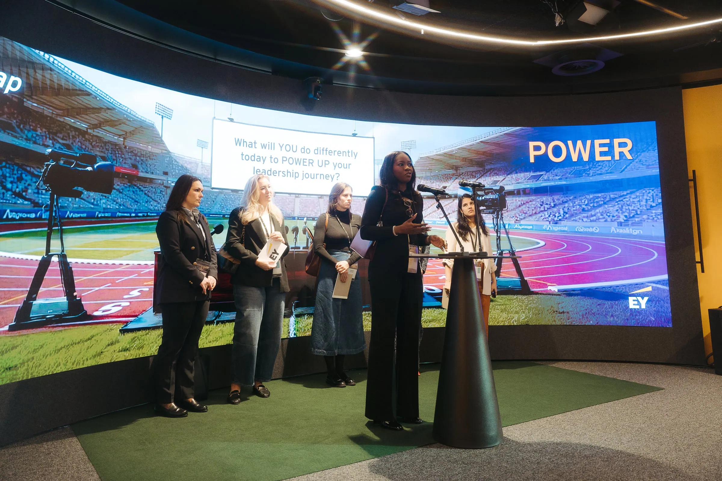 A woman speaking into microphones at a round lectern, with four other women and a large presentation screen behind her.