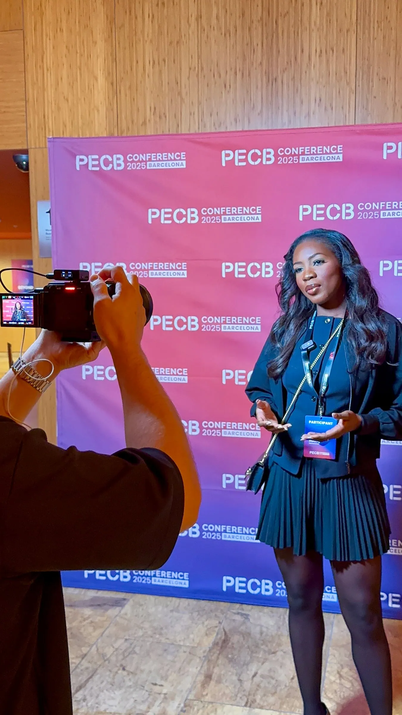 A woman in a dark blue outfit speaking and gesturing while being filmed in front of a pink backdrop.